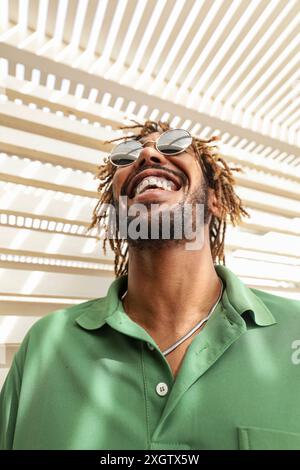 Un homme afro-américain avec des dreadlocks et des lunettes de soleil rit joyeusement, son visage relevé sous des ombres à motifs, vêtu d'une chemise verte. Banque D'Images