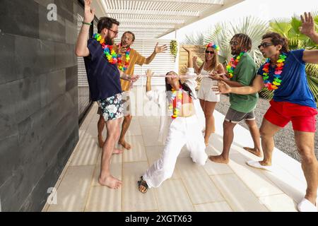 Un groupe dynamique d'amis danse joyeusement lors d'une fête estivale près d'une piscine, arbore un short décontracté et des leis fleuris, évoquant une ambiance tropicale festive. Banque D'Images