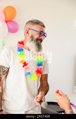 Un joyeux homme d'âge moyen lors d'une célébration festive, portant des lunettes bleues en forme de cœur et un lei vibrant, rit tout en tenant un micro, entouré Banque D'Images