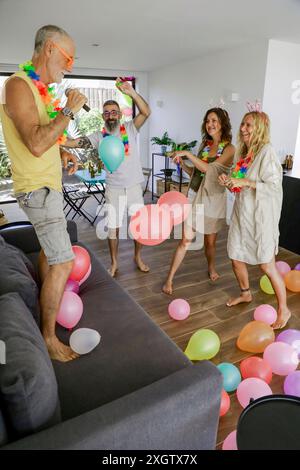 Des aînés joyeux dansant avec énergie dans un salon orné de ballons colorés, portant des chapeaux de fête et des leis tropicaux, célébrant la vie et les amis Banque D'Images