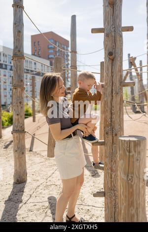 Une mère assiste son fils sur l'équipement d'un terrain de jeu dans un parc urbain. La mère et son fils regardent tous les deux vers le bas tout en s'engageant dans une activité ludique Banque D'Images