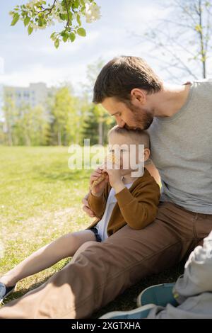 Dans cette scène estivale réconfortante, un père et son fils d'âge préscolaire sont assis sur l'herbe dans un parc, partageant un cône de crème glacée, père lui donne un doux kis Banque D'Images