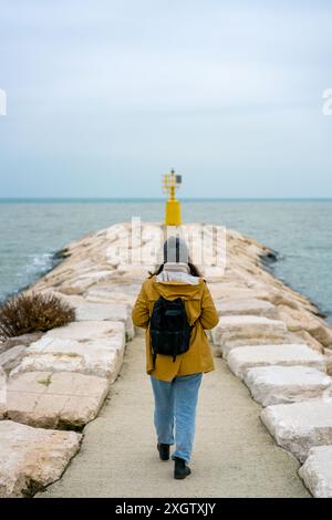 Une femme en veste jaune marche sur une jetée de pierre vers un phare jaune, naviguant sous un ciel nuageux par la mer tranquille à Rimini, Ital Banque D'Images