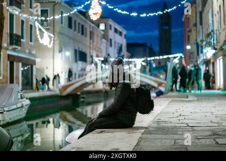 Une vue sereine sur un canal de Venise la nuit, avec une femme assise au bord de l'eau, avec des lumières de fête douces et des reflets Banque D'Images
