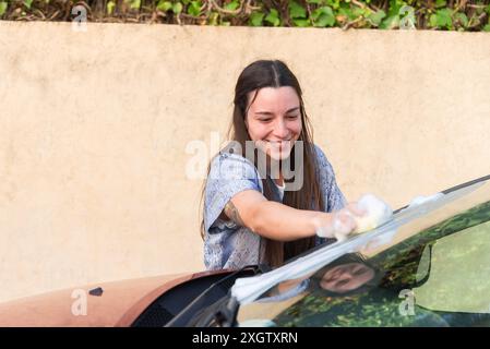 Une jeune femme joyeuse souriant alors qu'elle lave diligemment le pare-brise d'une voiture avec un chiffon Banque D'Images