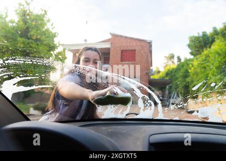 Une jeune femme joyeuse lave le pare-brise de sa voiture avec une éponge, souriant alors qu'elle travaille sous une toile de fond ensoleillée Banque D'Images