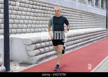 Un homme mûr aux cheveux gris, portant une chemise noire et un short, courant activement sur une piste de course rouge, avec un mur blanc structuré en arrière-plan Banque D'Images