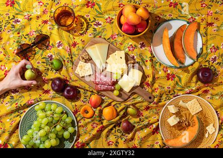 Vue aérienne d'une main anonyme dans un cadre de pique-nique d'été coloré, avec une planche de fromage en bois chargée de fromages variés, craquelins et salami, sur Banque D'Images