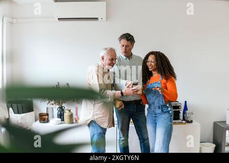 Trois collègues différents partagent du contenu sur un smartphone dans un espace de coworking lumineux et contemporain. La femme afro-américaine et ses deux colles caucasiennes Banque D'Images