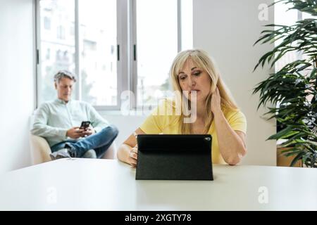 Une femme caucasienne regarde sa tablette dans un espace de coworking tandis qu'un homme caucasien utilise son smartphone en arrière-plan, illustrant ainsi le travail moderne Banque D'Images