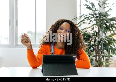 Femme afro-américaine regardant la caméra, souriante en travaillant sur une tablette dans un espace de coworking moderne. Elle porte une chemise orange et s'assoit dans une salle lumineuse Banque D'Images