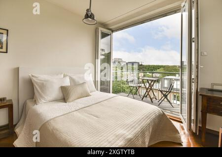 Une chambre accueillante dispose d''un lit confortable, d''une literie d''un blanc impeccable et d''un balcon meublé donnant sur un paysage urbain. Banque D'Images