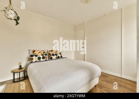Une chambre moderne confortable avec un lit blanc avec des draps rayés, des oreillers colorés, du parquet et un luminaire à plantes suspendues. Banque D'Images