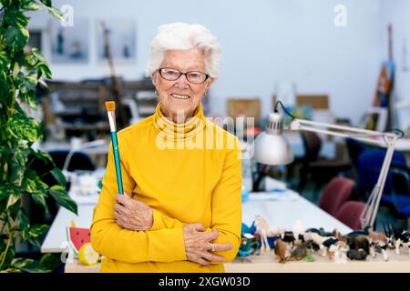 Une femme âgée joyeuse artiste aux cheveux blancs, portant un col roulé jaune, sourit tout en tenant un pinceau dans un studio d'art coloré Banque D'Images