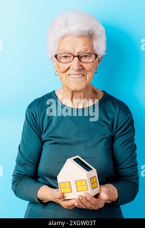 Une femme âgée joyeuse aux cheveux blancs, portant des lunettes et une chemise turquoise foncée, tient fièrement un petit modèle d'une maison équipée d'un panneau solaire, s. Banque D'Images