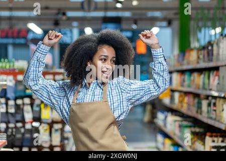 Joyeux employé de supermarché dans le tablier dansant et souriant dans l'allée du magasin. Fait preuve d'un environnement de travail positif, de bonheur, d'enthousiasme et de satisfaction au travail dans le secteur de la vente au détail Banque D'Images