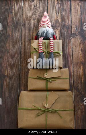 Trois cadeaux de Noël enveloppés dans du papier recyclé avec un gnome de Noël sur le dessus sur un fond en bois foncé. copier l'espace Banque D'Images
