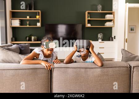 Adolescent biracial frère et sœur profiter des jeux VR à la maison. Ils s'amusent avec des casques de réalité virtuelle dans un cadre confortable de salon. Banque D'Images