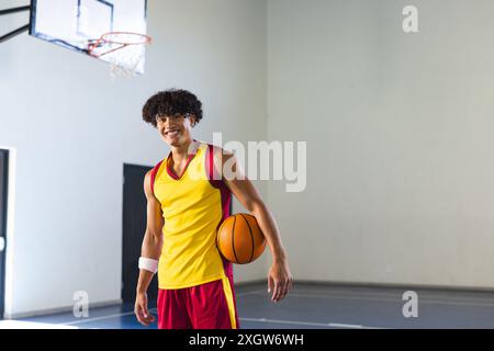 Jeune homme biracial pose dans un terrain de basket-ball, avec espace de copie. Son uniforme de basket-ball et son sourire éclatant véhiculent une passion pour le sport et un actif Banque D'Images