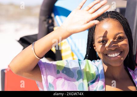 Jeune femme afro-américaine sourit brillamment à l'extérieur lors d'un Road trip. Son expression joyeuse et sa vague suggèrent une ambiance décontractée et ensoleillée de jour de plage. Banque D'Images