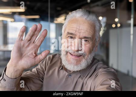 Homme âgé amical avec les cheveux gris et la barbe souriant et agitant à la caméra dans un environnement de bureau moderne, évoquant des sentiments de connexion, de convivialité et de chaleur. Banque D'Images