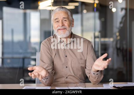 Homme d'affaires senior avec les cheveux gris et la barbe assis au bureau dans un environnement de bureau moderne exprimant des gestes avec les mains. Cadre professionnel avec lumière naturelle et murs de verre en arrière-plan. Banque D'Images