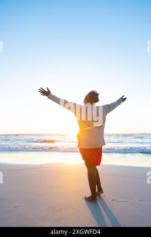 Une femme afro-américaine âgée profite d'un lever de soleil sur la plage. Elle embrasse le nouveau jour à bras ouverts, se sentant rafraîchie et paisible. Banque D'Images