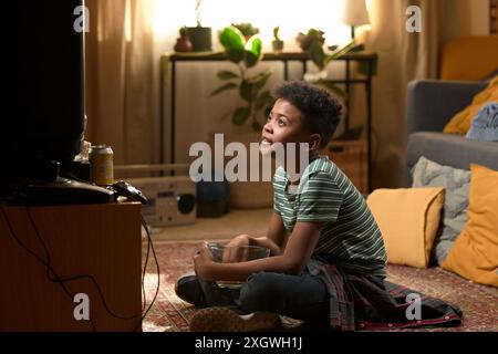 Écolier afro-américain assis les jambes croisées sur le sol regardant la télévision avec un bol de pop-corn sur ses genoux dans un salon confortable des années 80 Banque D'Images