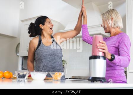Heureuses amies seniors diverses préparant un smoothie aux fruits avec un blender High Five dans la cuisine. Nourriture, nutrition, amitié, vie domestique et guérison Banque D'Images