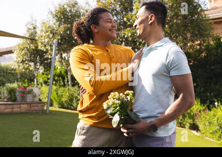 Jeune homme biracial et homme du moyen-Orient embrasser à l'extérieur, avec espace de copie. Ils partagent un moment joyeux dans un jardin, véhiculant un sentiment d'amour et de com Banque D'Images