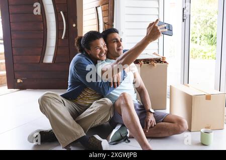 Un couple gay biracial et moyen-oriental prend un selfie, avec un espace de copie. Ils capturent un moment dans leur nouvelle maison, entourés de boîtes mobiles. Banque D'Images
