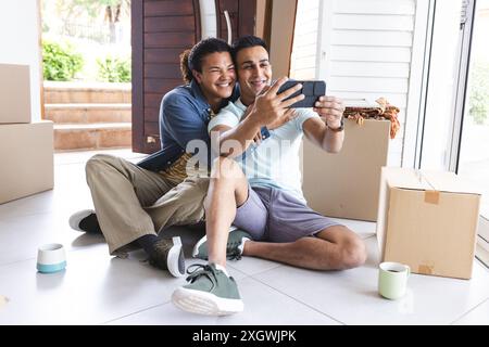 Couple gay biracial et moyen-oriental prendre un selfie à la maison. Ils partagent un moment joyeux lors d’une journée mouvante, entourés de boîtes. Banque D'Images