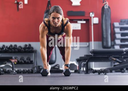 Fit jeune femme caucasienne effectue un exercice de planche au gymnase. Son expression ciblée souligne l'intensité de sa routine d'entraînement en force. Banque D'Images