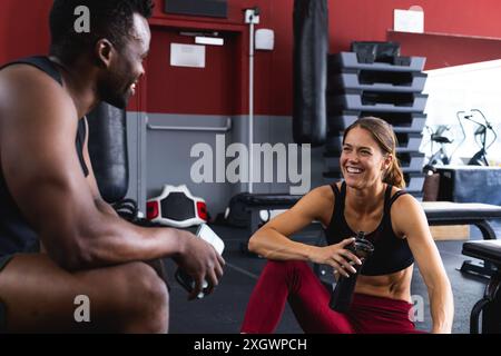 Fit homme afro-américain et jeune femme caucasienne partagent un rire au gymnase. Leur interaction amicale met en évidence l'aspect social de l'environnement de fitness Banque D'Images