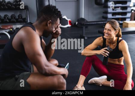 Fit homme afro-américain et jeune femme caucasienne partagent un rire au gymnase. Ils prennent une pause de leur entraînement, profitant d'un moment de camaradeur Banque D'Images
