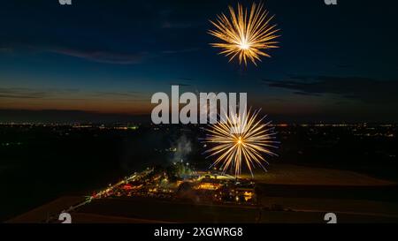 Deux feux d'artifice dorés explosent dans le ciel nocturne, créant des éclats brillants au-dessus d'Un rassemblement avec des tentes illuminées et les lumières lointaines de la ville, avec Un horizon crépusculaire ajoutant à la scène festive. Banque D'Images