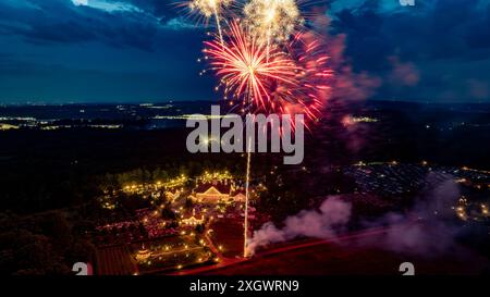 Feux d'artifice colorés explosant au-dessus d'Un domaine bien éclairé la nuit, entouré d'arbres et de champs, avec Un ciel vibrant et des lumières de ville lointaines sur l'horizon, capturant Une atmosphère festive de célébration. Banque D'Images