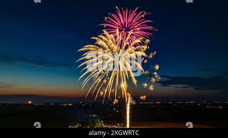 Feux d'artifice dorés et roses explosant dans le ciel nocturne, créant des éclats brillants sur le paysage urbain lointain et Une foule rassemblée, avec le vibrant Twilight Horizon rehaussant l'atmosphère festive. Banque D'Images