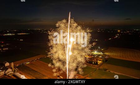 De brillants feux d'artifice dorés éclatent dans le ciel nocturne, créant Un spectacle éblouissant au-dessus d'Un parc d'expositions rural, illuminant les champs environnants et ajoutant Une lueur festive à l'ensemble de la scène. Banque D'Images