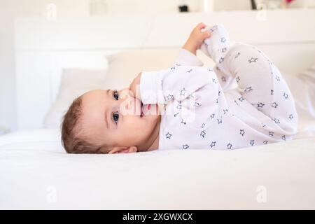 Adorable bébé en pyjama à motifs étoilés couché sur un lit blanc, souriant et ludique. Parfait pour les photos mettant en évidence la joie et l'innocence de l'enfance. Banque D'Images