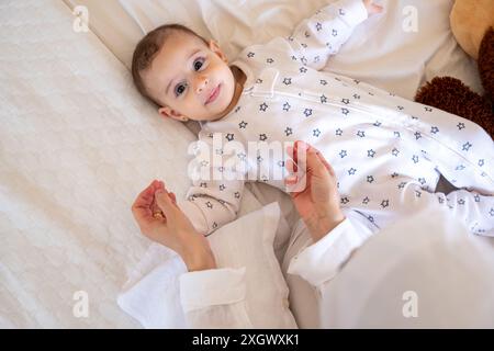 Bébé en pyjama à motifs étoilés couché sur un lit blanc avec un ours en peluche, assisté par un parent en hijab. Vue de dessus, gros plan, parfait pour la famille et les parents Banque D'Images