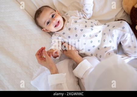 Bébé souriant en pyjama à motifs étoilés couché sur un lit blanc avec un ours en peluche, étant aimablement assisté par un parent. Idéal pour les photos de stock sur parentin Banque D'Images