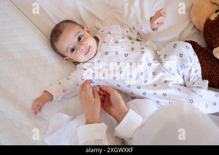 Bébé souriant dans un pyjama à motifs étoilés couché sur un lit blanc avec un ours en peluche, étant amoureusement assisté par un parent. Idéal pour les photos de stock sur parentin Banque D'Images