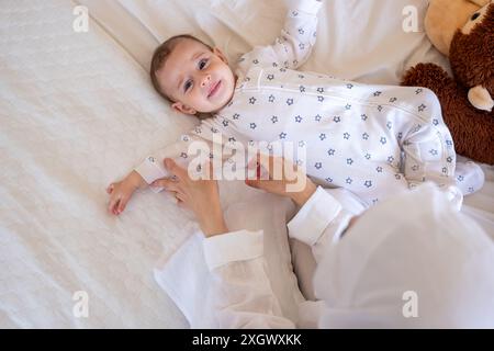 Bébé souriant en pyjama à motifs étoilés couché sur un lit blanc avec un ours en peluche, étant aimablement assisté par un parent. Idéal pour les photos de stock sur parentin Banque D'Images