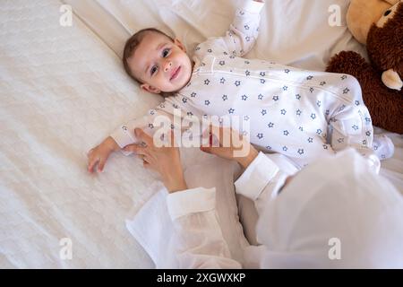 Bébé souriant en pyjama à motifs étoilés couché sur un lit blanc avec un ours en peluche, étant aimablement assisté par un parent. Idéal pour les photos de stock sur parentin Banque D'Images