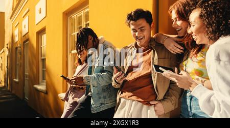 Un groupe d'amis heureux partageant des moments sur leur smartphone contre un mur jaune vif à l'extérieur par une journée ensoleillée. Banque D'Images