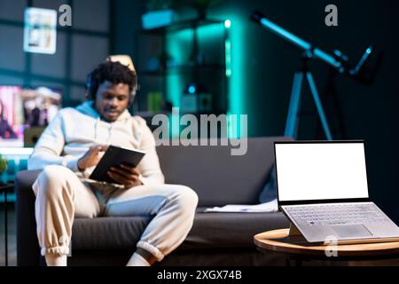 Homme assis sur le canapé, lisant eBook à partir d'une tablette dans l'appartement, concentrez-vous sur un ordinateur portable maquette. Personne détendue portant des écouteurs, écoutant un livre audio à côté d'un dispositif d'écran isolé sur une table basse Banque D'Images