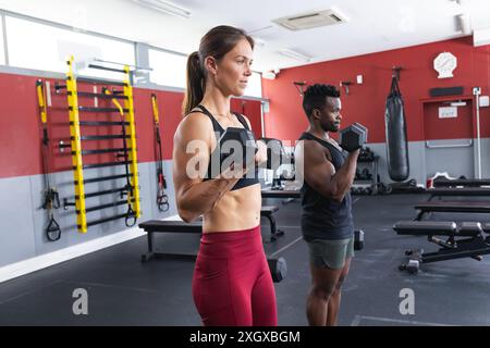 Fit jeune femme caucasienne et homme afro-américain soulevant des poids au gymnase. Leur routine d'entraînement ciblée met en valeur la force et la détermination. Banque D'Images