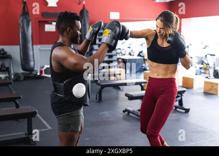 Fit homme afro-américain entraînerait jeune femme caucasienne fit dans la boxe au gymnase. Ils se concentrent sur la technique lors d'une séance d'entraînement personnelle. Banque D'Images