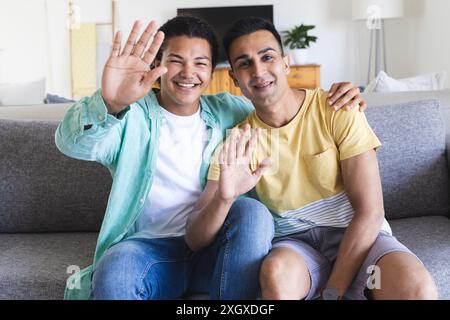 Couple gay diversifié assis sur un canapé à la maison pendant un appel vidéo, avec espace de copie. Ils partagent un moment joyeux, agitant à la caméra avec Bright Banque D'Images